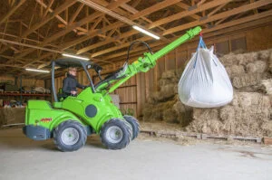 A green Avant 528 mini front loader lifting a large bag of materials inside a barn. The machine is equipped with a telescopic boom and operated by a person seated in the cabin. The surrounding area includes stacks of hay bales, wooden beams, and a storage structure in the background.